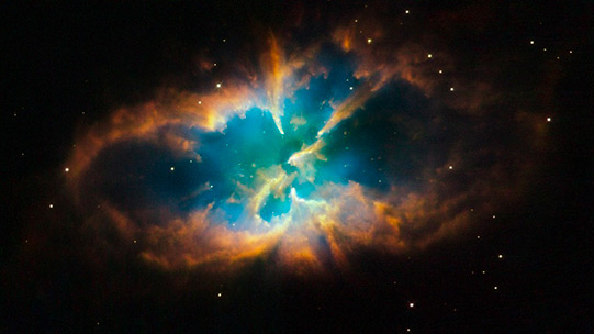 This planetary nebula, known as NGC 2818, takes up the entire frame. It is surrounded by an orange oval of wispy clouds shaped almost like lips. Inside the oval is a bright blue cloud pierced by a few small spikes of the orange clouds. 